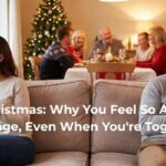 A wide-angle photograph illustrating marital loneliness during the holidays. A couple sits at opposite ends of a sofa; the wife gazes sadly out a window while the husband is engrossed in his phone. In the background, a happy multi-generational family enjoys Christmas dinner near a decorated tree, creating a sharp contrast to the couple's isolation.