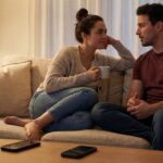 A couple sits together on a couch, practicing active listening by making eye contact and having a focused conversation. Their phones are placed face down on the coffee table, symbolizing a commitment to disconnect from distractions and improve communication in their relationship.