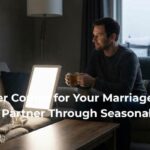 A supportive husband sits with his wife who is wrapped in a blanket and using a light therapy lamp for Seasonal Affective Disorder (SAD) on a snowy winter day.