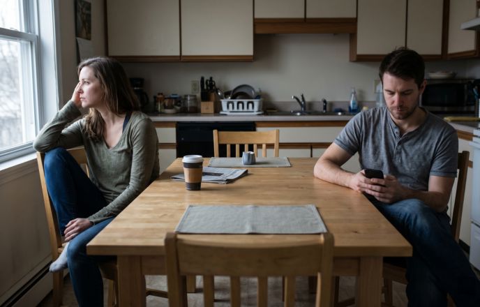Two people sitting apart at a kitchen table, each looking away, illustrating the emotional distance that AI tools can help couples begin to bridge before a real conversation